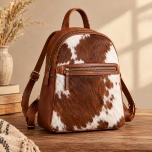 Puede incluir: Una mochila de piel de vaca en color marrón y blanco con asa superior, correas ajustables y un bolsillo frontal con cremallera. El cuerpo principal muestra un patrón natural de manchas con ribetes de cuero liso y herrajes de latón.