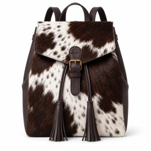Puede incluir: Una mochila con un patrón de piel de vaca en color marrón oscuro y blanco en la solapa frontal y el cuerpo. La bolsa incluye correas de color marrón oscuro, un asa superior, un cierre central con hebilla de metal y dos borlas largas.