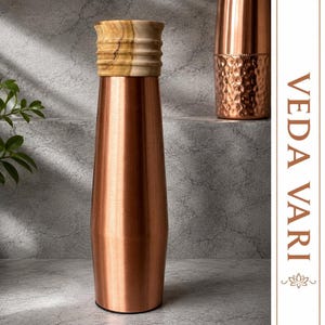 Puede incluir: Una botella de agua de cobre lisa y alta con cuerpo cónico y tapa de madera estriada de color claro. El texto VEDA VARI está impreso verticalmente en una fuente de color cobre en el lado derecho, acompañado de un pequeño icono de loto.