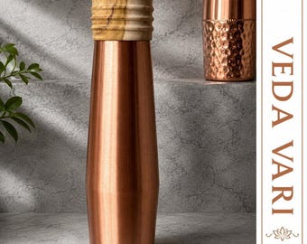 Puede incluir: Una botella de agua de cobre lisa y alta con cuerpo cónico y tapa de madera estriada de color claro. El texto VEDA VARI está impreso verticalmente en una fuente de color cobre en el lado derecho, acompañado de un pequeño icono de loto.