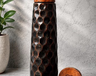 Puede incluir: Una botella de agua de metal marrón oscuro con textura martillada, forma cilíndrica y una tapa de madera marrón claro. Una tapa de madera a juego se encuentra al lado de la botella, con un logotipo circular con las letras AC grabadas.