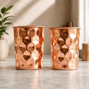 Puede incluir: Dos vasos cilíndricos de cobre con un patrón martillado repetitivo de hendiduras redondeadas y entrelazadas en la superficie exterior. El metal tiene un acabado pulido y reflectante que capta la luz, creando brillos y sombras.