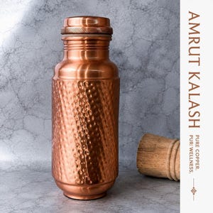 May include: A copper water bottle with a hammered texture on the body and a smooth neck and cap. The text AMRUT KALASH, PURE COPPER, and PURI WELLNESS is printed on the right side. A wooden stopper is positioned to the right of the bottle.