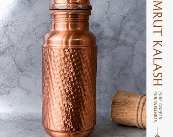 Puede incluir: Una botella de agua de cobre con textura martillada en el cuerpo y cuello y tapa lisos. El texto AMRUT KALASH, PURE COPPER y PURI WELLNESS está impreso en el lado derecho. Un tapón de madera está colocado a la derecha de la botella.