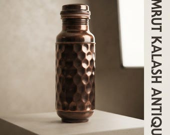 Puede incluir: Una botella de agua cilíndrica de cobre con un acabado oscuro martillado y una tapa de rosca a juego. La botella presenta una etiqueta de texto vertical en el lado derecho que dice AMRUT KALASH ANTIQUE en letras mayúsculas negras.