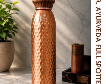 Puede incluir: Una botella de agua cilíndrica de cobre con una textura martillada y una tapa de madera marrón oscuro. El texto DR. AYURVEDA FULL SPOTTED está impreso verticalmente a lo largo del lado derecho del marco de la imagen.