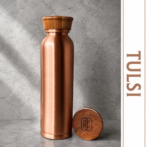 Puede incluir: Una botella de agua cilíndrica de cobre con un acabado metálico liso y una tapa de madera oscura. Una tapa circular de madera a juego descansa junto a la botella, con un logotipo negro con las letras AC. La palabra TULSI está impresa verticalmente en marrón.