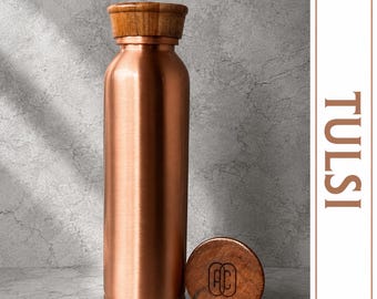 May include: A cylindrical copper water bottle with a smooth metallic finish and a wooden cap. A matching wooden cap sits beside the bottle, featuring the letters AC engraved in the center. The word TULSI is printed vertically on the right side.