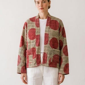 Könnte beinhalten: Eine langärmelige, vorne offene Jacke mit beigefarbener Basis, einem sich wiederholenden dunkelgrünen Fischgrätenmuster und großen, einfarbigen roten Kreismotiven. Das Kleidungsstück hat zwei aufgesetzte Vordertaschen und einen Schalkragen.