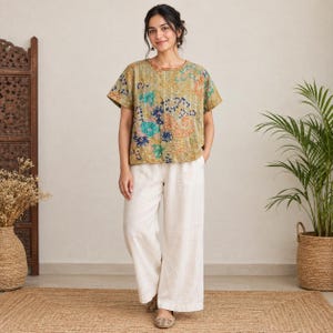 Könnte beinhalten: Ein kurzärmeliges Oberteil in gedämpftem Olivgrün mit einem mehrfarbigen Blumen- und Paisley-Muster sowie sichtbaren Steppnähten, kombiniert mit einer weit geschnittenen, bodenlangen Hose in Off-White mit lockerer Passform.