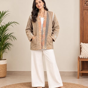 May include: A long-sleeved, open-front jacket in a muted tan and beige tone with a textured, quilted appearance. The jacket features a contrasting vertical panel in shades of orange and grey along the front opening, paired with white wide-leg trousers.