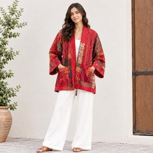 Può includere: Una giacca patchwork rossa con una trama trapuntata e cuciture visibili, maniche lunghe e due tasche frontali. La giacca è indossata sopra un top bianco semplice e pantaloni bianchi a gamba larga, abbinata a sandali marroni aperti.