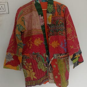 May include: A long-sleeve jacket constructed from patchwork fabric featuring a vibrant mix of red, orange, green, and yellow colours. The garment displays various floral and geometric patterns with visible white running stitch embroidery throughout.