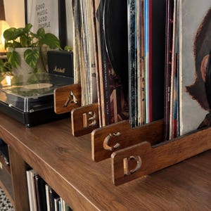 May include: A set of four wooden vinyl record dividers, each featuring a cutout letter of the alphabet, specifically A, B, C, and D. These rectangular dividers are positioned upright to organise a collection of vinyl records on a wooden surface.