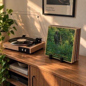 May include: A light wood turntable with a clear dust cover and a black platter, positioned next to a stack of vinyl records in a black stand. The front record features a green forest scene with a person playing a saxophone and Japanese text.