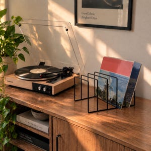 May include: A light wood turntable with a clear dust cover and a black metal wire record storage rack holding two vinyl records. The turntable features a black control panel with two silver knobs and the brand name Fluance printed on the front.