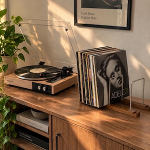May include: A wooden record player with a clear plastic dust cover and a black turntable platter. Next to it, a wooden record holder displays a collection of vinyl records, including the Adele 21 album cover, arranged in a vertical metal rack.