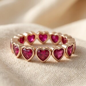 May include: A gold-toned eternity ring featuring a continuous band of heart-shaped, vibrant pink-red gemstones. Each stone is individually set within a polished metal frame, creating a repeating heart pattern that encircles the entire finger band.