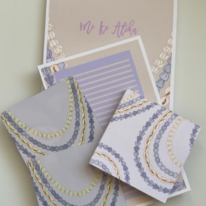 Könnte beinhalten: Ein Schreibwarenset bestehend aus einem beigen Blatt mit dem Text Me ke Aloha, einer lila linierten Notizkarte und zwei quadratischen Karten, alle verziert mit Illustrationen von geschichteten Muschel- und Blumenketten in Lila, Gelb und Weiß.