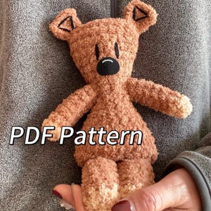 May include: A brown plush toy with a fuzzy texture, featuring embroidered black triangular ear details, two vertical black lines for eyes, and a black oval nose with a curved mouth. The text PDF Pattern is printed in white over the toy.
