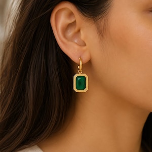 May include: A gold hoop earring featuring a rectangular pendant with a faceted deep green stone. The pendant has a textured, beaded border surrounding the central gemstone, which is suspended from a simple polished gold circular hoop earring.