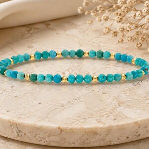 May include: A beaded bracelet featuring small, round turquoise-coloured stones in various shades of blue and green, interspersed with tiny, polished gold-toned metal beads arranged in a repeating pattern along the entire length of the circular band.