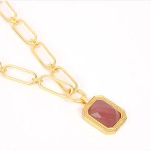 Puede incluir: Un collar de cadena en tono dorado con eslabones ovalados alargados y un colgante rectangular. El colgante tiene un borde dorado texturizado y una piedra facetada de color rosa empolvado en el centro, creando un aspecto pulido.