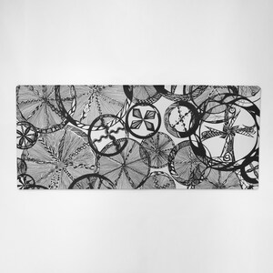 Pode incluir: Um tapete de secretária retangular com uma ilustração a tinta preta e branca de padrões circulares sobrepostos. O design consiste em várias formas geométricas complexas, linhas finas e texturas de hachuras contidas em vários círculos.
