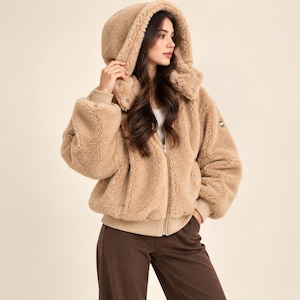 Puede incluir: Una chaqueta de piel sintética beige con capucha, cremallera frontal completa, mangas largas con puños acanalados y dobladillo acanalado. La chaqueta presenta un material grueso y texturizado, una capucha grande y un pequeño parche con logo.