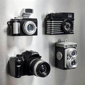 Peut inclure: Un ensemble de quatre aimants en forme d'appareils photo. L'un a un flash argenté, un autre des rayures noires et blanches avec le texte Glass lens 5.0 Megapixel, un troisième est un reflex noir avec Camera, et le quatrième est un bi-objectif Rock n Roll.