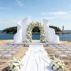 May include: A wedding ceremony setup on a sandy beach featuring a large white floral archway flanked by two tall, curved white fabric panels. A long white fabric aisle runner leads to the arch, surrounded by rows of clear acrylic chairs.