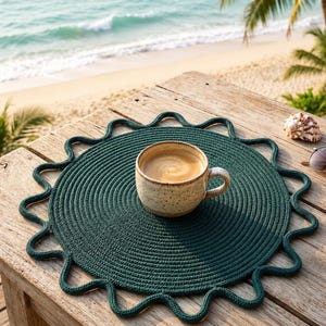May include: A round, dark green woven placemat with a scalloped, wavy edge design. Centered on the placemat is a speckled, light-coloured ceramic mug filled with a coffee beverage. The placemat features a tight, circular rope-like texture.