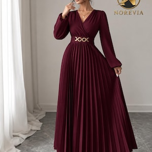 May include: A floor-length burgundy dress featuring long sleeves, a V-neckline, and a pleated skirt. The waist is accented with a gold-toned metal link belt. The brand logo NOREVIA is visible in the upper right corner of the image.