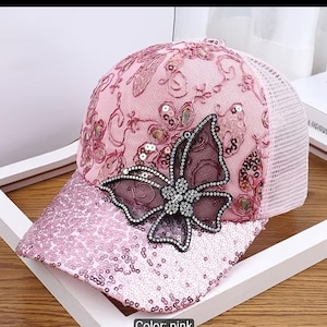 Peut inclure: Une casquette de baseball rose avec un arrière en filet, une visière à paillettes et une application papillon sur le devant. La calotte est ornée de broderies florales et de paillettes, tandis que le papillon est bordé de petites perles argentées.
