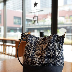 Könnte beinhalten: Eine dunkelblaue Beuteltasche mit einem weißen, gestickten Gittermuster aus Schneeflocken und Schmetterlingen. Die Tasche hat einen einfarbigen, dunkelblauen, strukturierten Boden und einen dünnen, dunkelblauen Schulterriemen mit goldenen Metallringen.