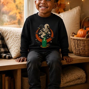 May include: A black long-sleeve sweatshirt featuring a graphic print of a jester character emerging from a green candle flame, set against an orange crescent moon with small stars and bats, paired with dark gray denim jeans and black sneakers.