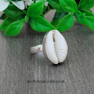 Può includere: Un anello in argento con una conchiglia di ciprea bianca come elemento centrale. La conchiglia ha una forma ovale liscia con una fessura centrale e una consistenza striata. La fascia in metallo è lucida e semplice. Il testo BHERUBABAJEWELSHUB è visibile in basso.