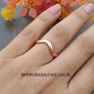 Könnte beinhalten: Ein glatter, polierter Ring aus Roségold mit einem markanten, wellenförmigen Band. Der Ring weist ein durchgehendes, geschwungenes Design auf, das einem sanften Wellenmuster folgt. Der Text BHERUBABAJEWELSHUB ist schwarz im unteren Bildbereich gedruckt.