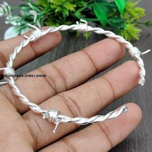Può includere: Un bracciale rigido color argento progettato per assomigliare a filo spinato intrecciato con piccole punte metalliche avvolte a intervalli lungo la fascia. Il testo BHERUBABAJEWELSHUB è stampato in nero sul lato sinistro del bracciale.