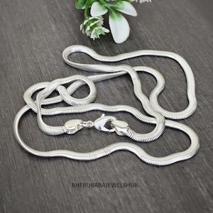 Puede incluir: Un collar de cadena de serpiente en tono plateado con un diseño flexible, liso y de eslabones planos. El collar incluye un cierre de mosquetón y presenta el texto BHERUBABAJEWELSHUB en la parte inferior de la imagen.