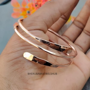 Può includere: Un bracciale a spirale color oro rosa caratterizzato da una texture martellata e lucida sulle estremità affusolate. La fascia metallica si avvolge in un design a doppio giro. Il testo BHERUBABAJEWELSHUB è stampato in nero in basso nell'immagine.