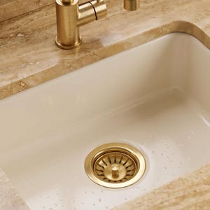 Könnte beinhalten: Ein rechteckiges, cremeweißes Küchenspülbecken mit einer Armatur in gebürstetem Gold und einem passenden goldenen Siebkorb. Das Becken ist in eine polierte, beige Travertin-Arbeitsplatte mit natürlicher Maserung und dunkelgrünem Unterschrank eingelassen.