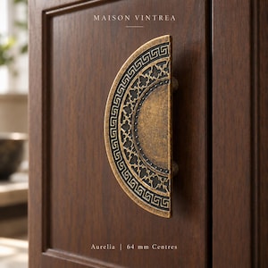 Puede incluir: Un tirador de armario semicircular en tono latón con un borde griego oscuro y hundido y un patrón central de cuatrifolio. El herraje está montado en una puerta de armario de madera oscura. El texto dice MAISON VINTREA, Aurelia, 64 mm Centres.
