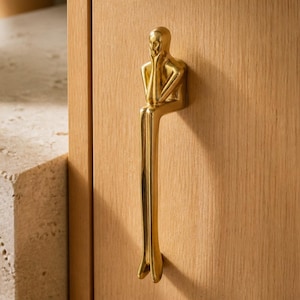 May include: A polished gold-toned metal cabinet handle shaped like a stylized human figure sitting with legs extended downward. The figure rests its chin on its hands, with a smooth, reflective surface mounted vertically onto a light wood panel.