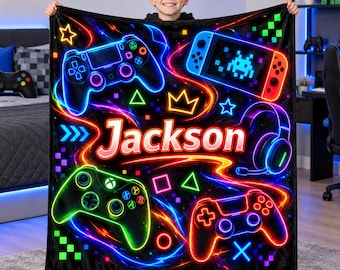 May include: A black fleece blanket featuring a vibrant neon-style print of various video game controllers, a handheld console, and a headset. The name Jackson is printed in large, glowing white letters in the center, surrounded by colorful light trails.