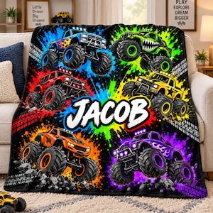 May include: A black fleece blanket featuring five colorful monster trucks in blue, green, red, yellow, orange, and purple, surrounded by vibrant paint splatters and tire tracks. The name JACOB is printed in large white letters in the center.
