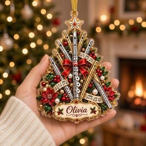 Puede incluir: Un adorno con forma de árbol de Navidad que presenta flautas plateadas y doradas en abanico, decorado con lazos rojos, bayas de acebo, perlas blancas y notas musicales. Una placa central en la base muestra el nombre Olivia en cursiva.