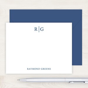 May include: A white stationery card featuring a dark blue monogram with the letters R and G separated by a vertical line, and the name RAYMOND GREENE printed in dark blue at the bottom, accompanied by a matching dark blue envelope and a silver pen.