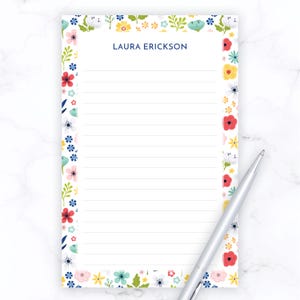 May include: A lined notepad with a floral border featuring red, yellow, blue, and pink flowers. The notepad has the name "LAURA ERICKSON" printed at the top. A silver pen rests on the notepad.