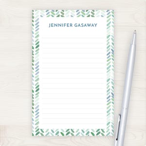 May include: A rectangular notepad with a white background and lined pages. The notepad has a border of green and blue leaf-like shapes. The name "JENNIFER GASAWAY" is printed at the top. A silver pen rests on the right side of the notepad.